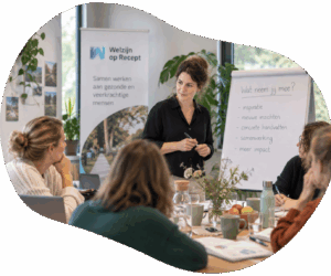 Trainingssituatie. Meerdere deelnemers zitten aan een houten tafel met daarop een bosje bloemen, diverse boeken, een schaaltje fruit en bekers met drinken. Aan het hoofd van de tafel staat een vrouwelijke trainer. Zij heeft donderkbruin haar in een loss knot en een zware blouse aan. Een pen in de handen en kijkt een deelnemer aan. Op de achtergrond zijn een flip-over met het logo van Welzijn o Recept te zien en een flipover met aantekeningen. Daarachter zijn ramen en planten te zien.